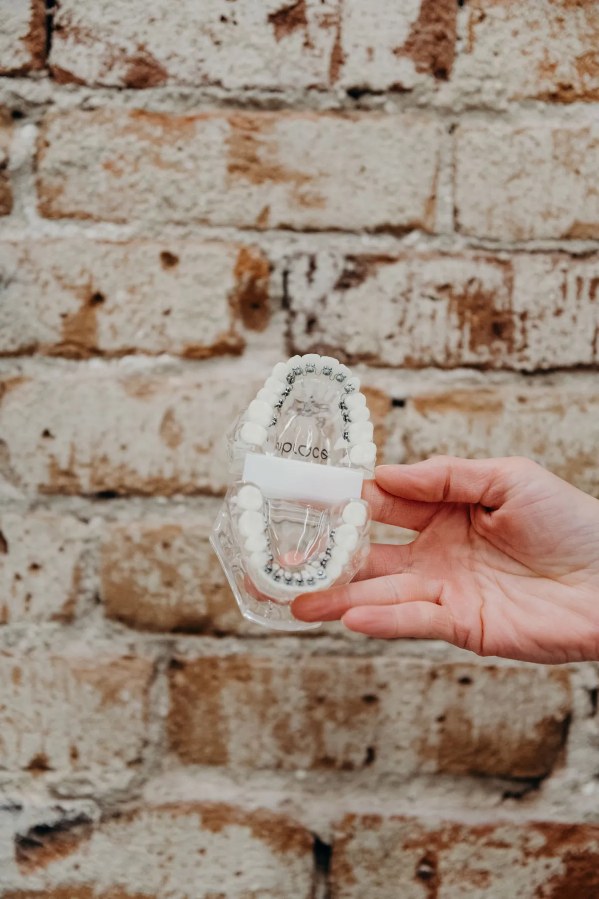 Hand holding a clear teeth model with lingual braces in front of a brick wall at aligned. Orthodontics